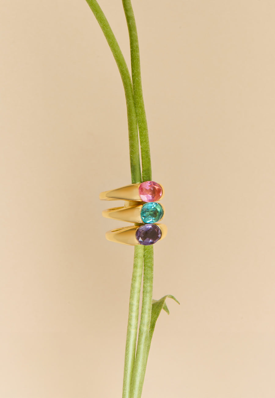 Tres anillos dorados The Empress de Lavani Jewels con piedras ovaladas en rosa, azul y violeta abrazan delicadamente un tallo floral en una composición elegante y sofisticada.