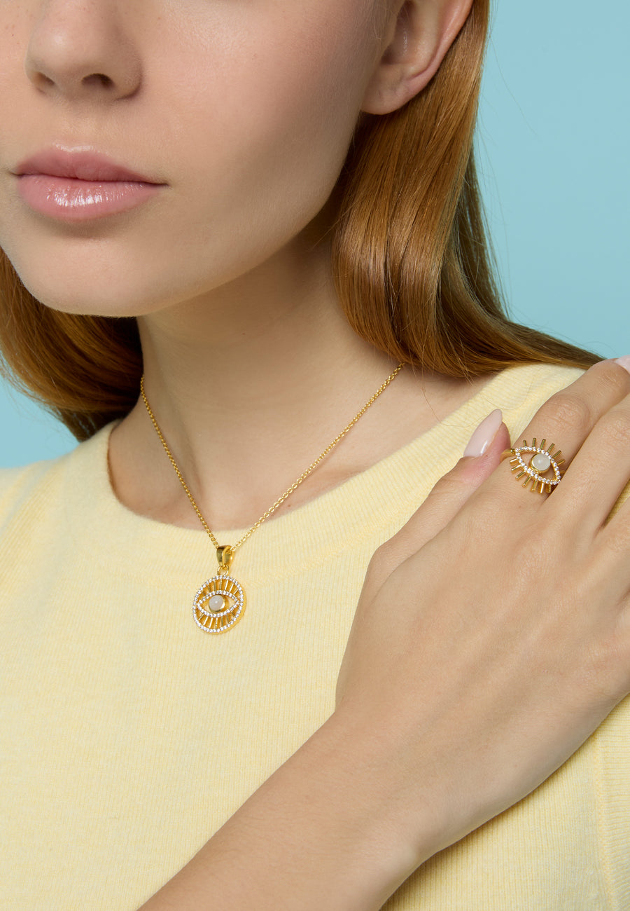 Mujer luciendo un anillo dorado con diseño de ojo turco acompañado de un collar a juego de Lavani Jewels sobre fondo azul claro.