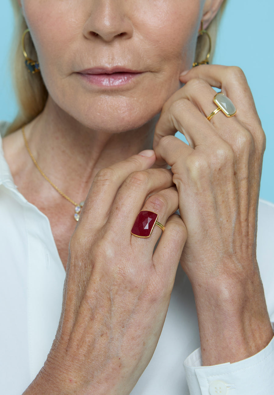 Mujer mayor con camisa blanca mostrando dos anillos Stardust de Lavani Jewels, uno con piedra aguamarina y otro con rubí, acompañados por un collar delicado y pendientes dorados, sobre fondo azul claro.