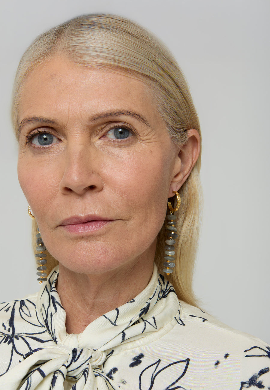 Mujer rubia con blusa blanca estampada luciendo los Aros Mizu en tono gris de Lavani Jewels, pendientes dorados con cuentas apiladas de piedra gris.