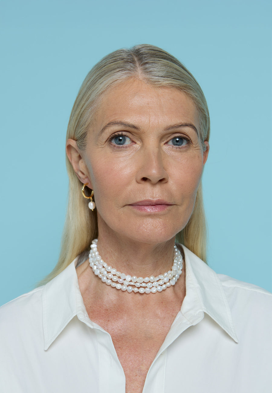Mujer de cabello rubio y camisa blanca luciendo el choker Mizu de perlas de Lavani Jewels junto a un pendiente dorado con perla colgante, sobre fondo azul claro.