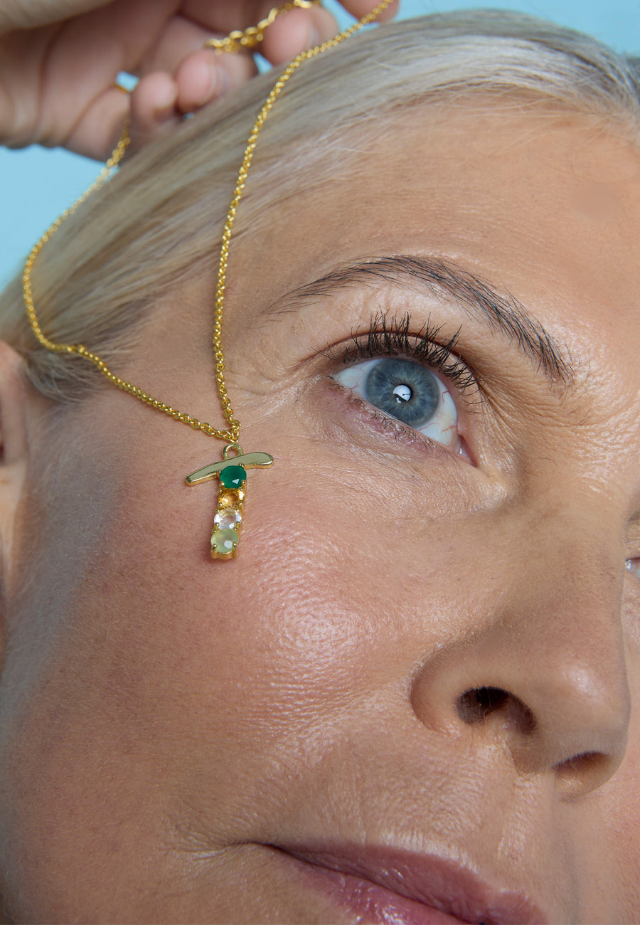 Collar dorado con colgante de inicial T y piedras de colores de Lavani Jewels, sostenido sobre el rostro de una mujer rubia de ojos azules.