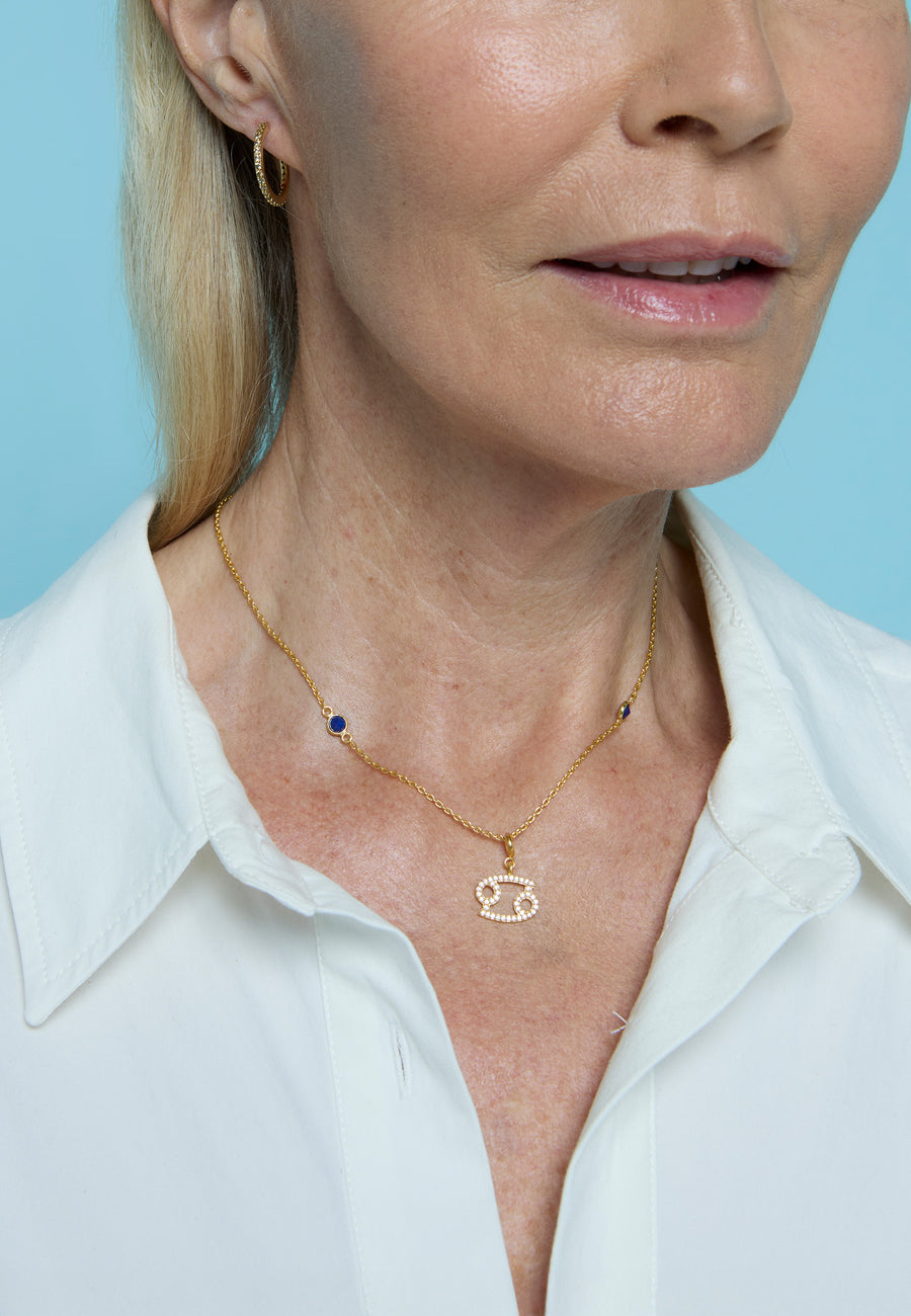Mujer rubia con camisa blanca luciendo el collar de signo zodiacal Cáncer de Lavani Jewels, con charm dorado y circonitas, sobre fondo azul claro.