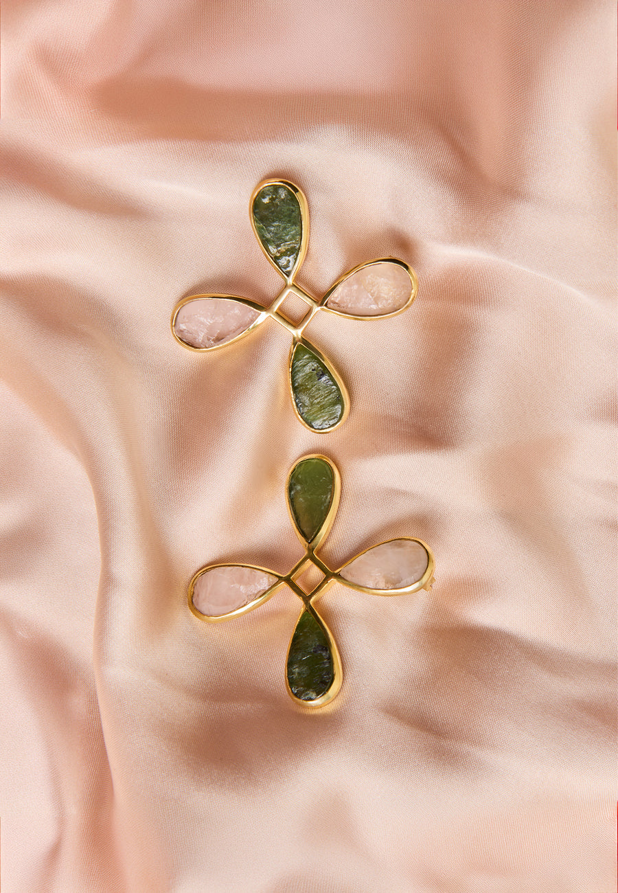 Pendientes Cruz Namasté de Lavani Jewels con piedras de cuarzo rosa y verde, colocados sobre un fondo de raso rosa suave.
