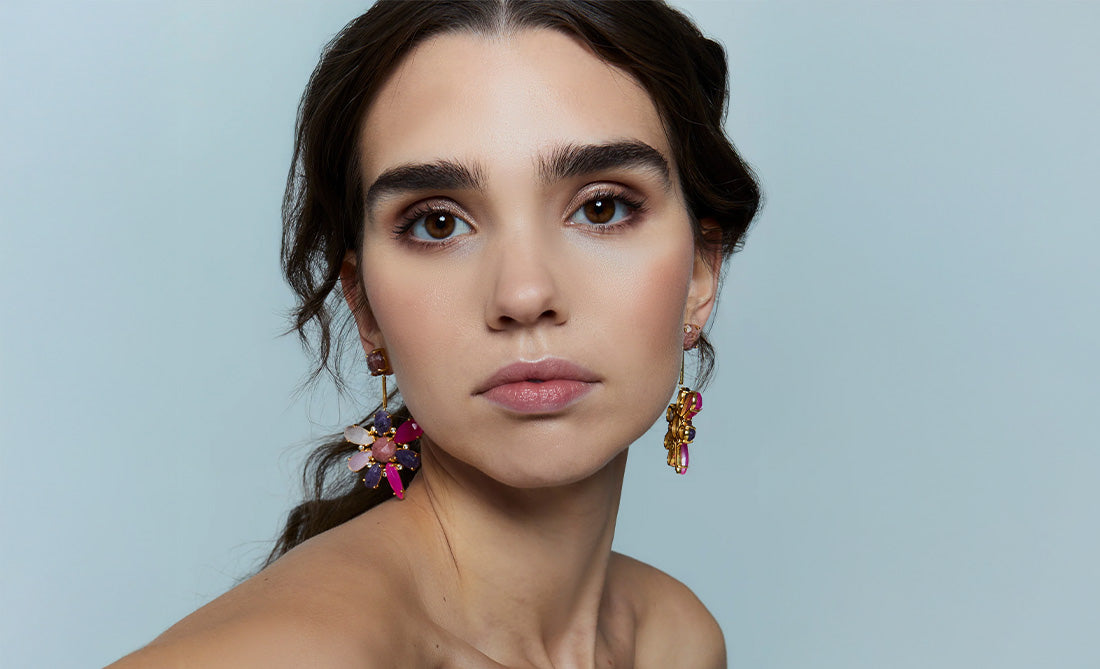 Mujer con pendientes dorados tipo flor en tonos buganvilla y rosa, con fondo celeste.