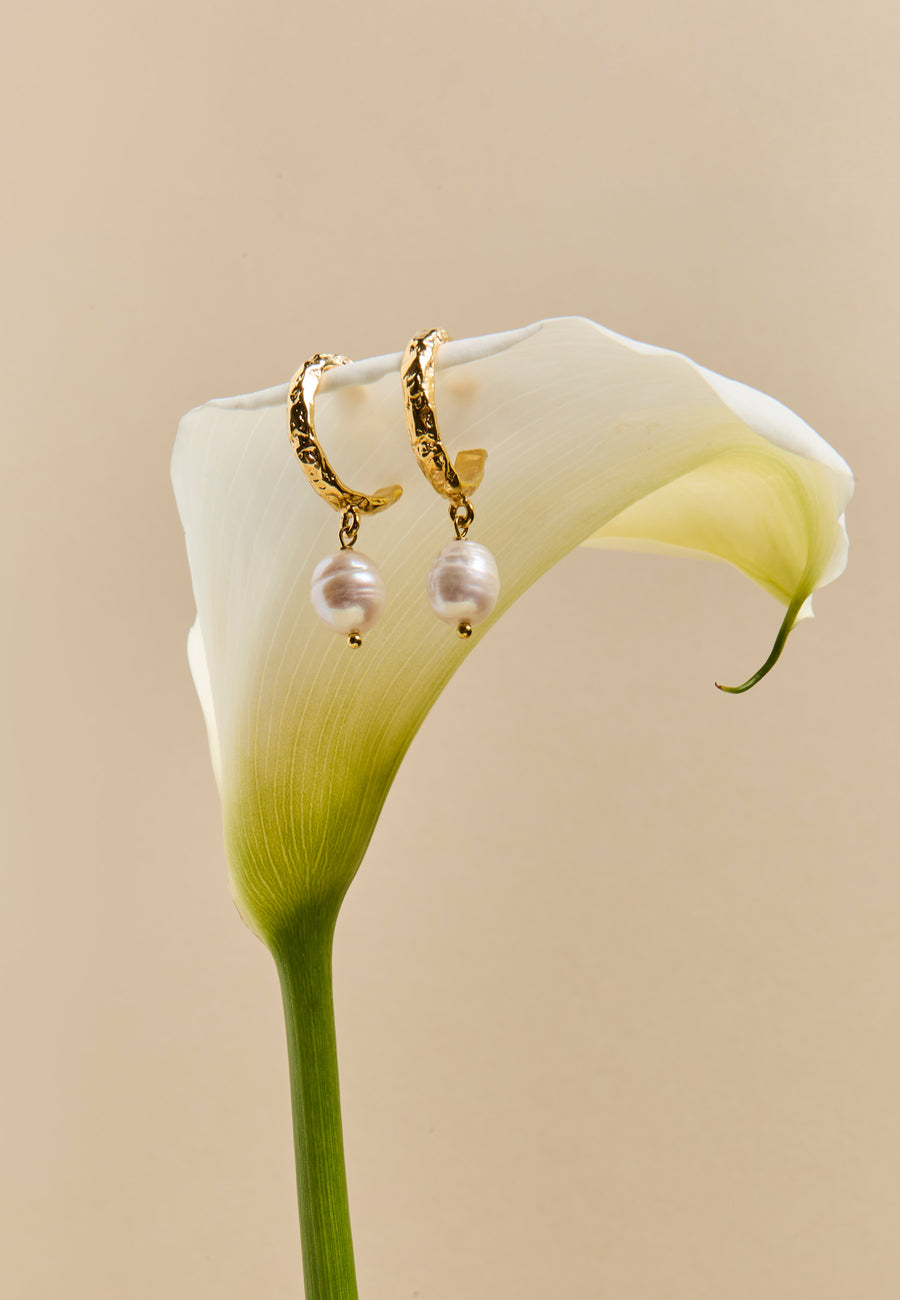 Pendientes dorados con perla natural inspirados en la piedra birthstone de Abril, colgados sobre flor de cala blanca, joya sofisticada para novia o evento especial.