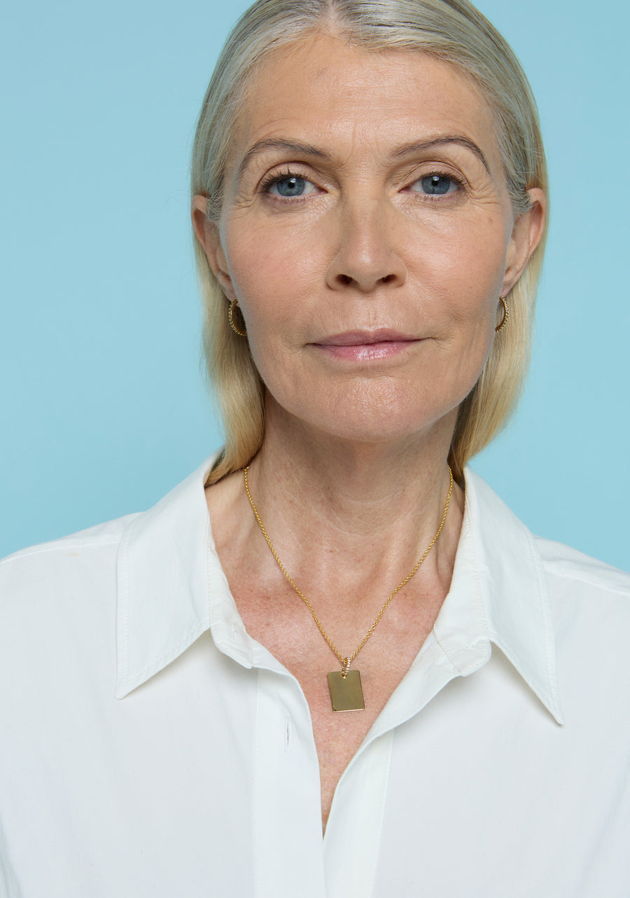 Mujer rubia con camisa blanca luciendo el collar Signature de Lavani Jewels, con colgante rectangular dorado grabado, sobre fondo azul claro.