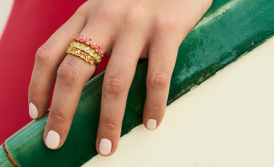 Mano con anillos dorados texturizados con piedras naturales en tonos rosa, amarillo y blanco, joyería apilable colorida.