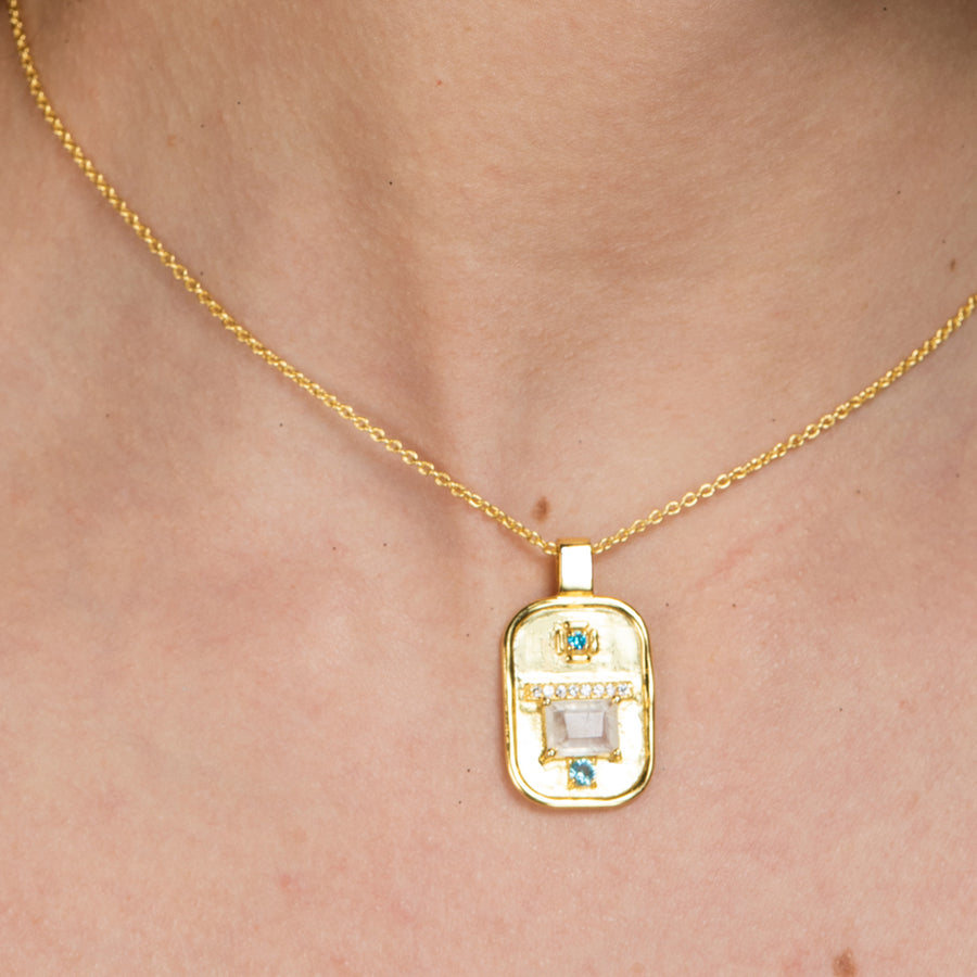 Mujer porta colgante mediano con medalla rectangular sujeta por una fina cadena dorada y adornada con piedras naturales.